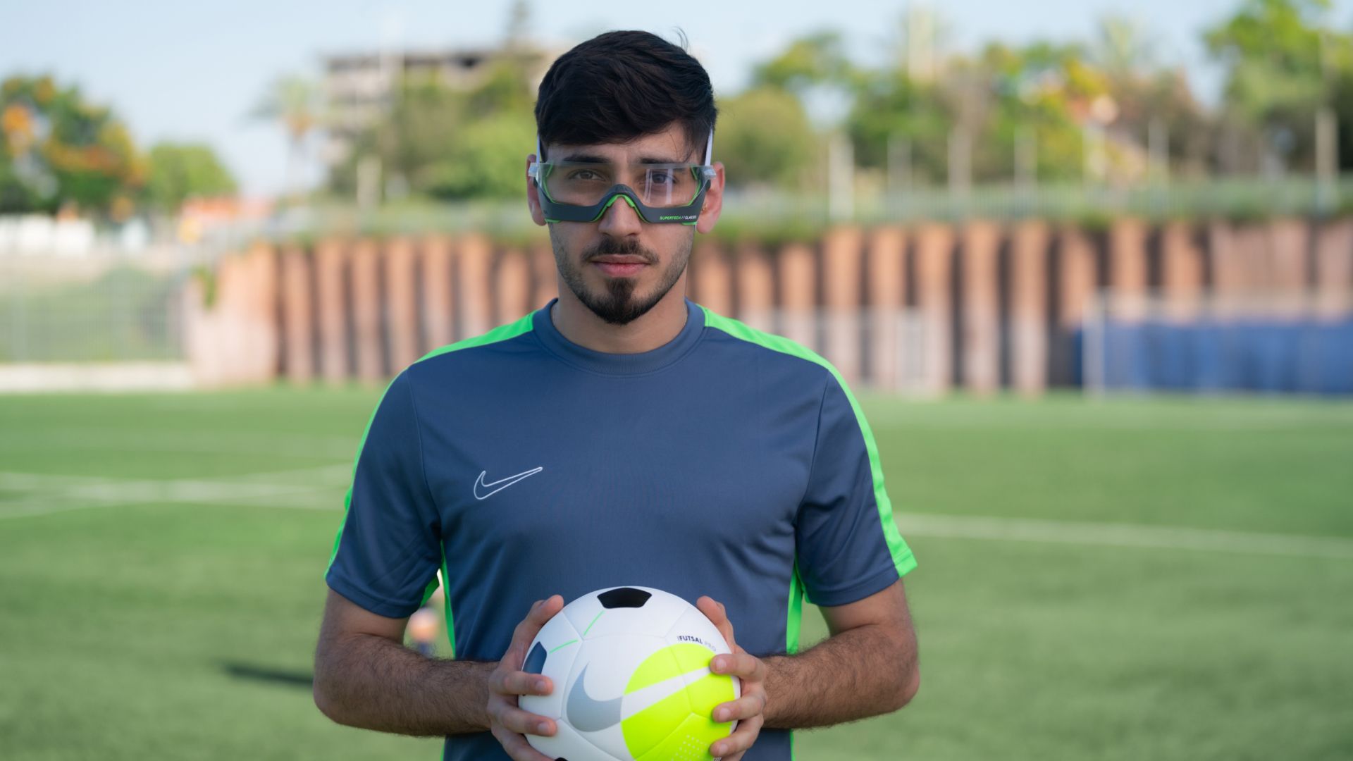 A young footballer standing on the pitch holding a ball, wearing the Rayzor glasses.